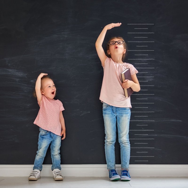 Two sisters measuring their growth against a chalkboard height chart